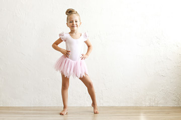 Little blonde balerina girl dancing and posing in dance club with wooden floot an white textured plaster wall. Young ballet dancer in pink tutu dress, having fun and smiling. Backgroud, copy space.