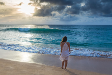 Young woman in white dress walking on the beach at sunset and staring at the sun, Oahu North shore, Hawaii