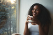 © Anatoliy Karlyuk - Close up view of fashionable cute young African American female in white tank top having rest indoors, holding large cup of hot tea, smiling broadly, daydreaming, spending nice time at home alone