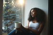 © Anatoliy Karlyuk - Portrait of beautiful twenty year old African American female wearing tank top and ripped jeans enjoying urban view while sitting on windowsill with mug of hot drink, feeling cozy and relaxed