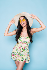 Poster - Portrait of cute summer brunette woman in hat, sunglasses and colorful dress, stylish girl have a fun and posing on blue background