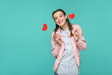 Wall Mural - Happy beautiful girl in casual style, pigtail hairstyle and pink jacket, standing and holding red heart stickers and looking at camera and toothy smiling, Indoor, isolated on blue or green background