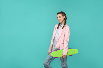 Wall Mural - happy beautiful girl in casual style, pigtail hair and pink jacket, standing and holding skateboard and looking at camera with toothy smile, Indoor isolated, studio shot on blue or green background