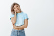 © Cookie Studio - Indoor shot of feminine charming woman in stylish clothes, tilting head and smiling while touching neck and holding arm crossed over chest, being timid and unconfident standing in awkward situation