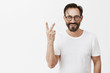 Indoor shot of beautiful confident and happy man with trendy glasses showing number two or up yours gesture, smiling broadly over gray background, making something twice times