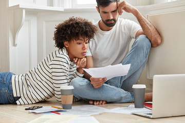 Young mixed race family couple try to solve financial situation, study business papers, plan budget, make schemes on laptop computer, drink coffee, pose on floor in living room with light colours