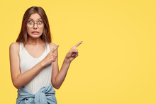 Oops, Come And Look There! Lovely Young Woman Feels Worried And Ashamed, Points With Both Index Fingers At Upper Right Corner, Isolated Over Yellow Background, Makes Mistake And Looks Awkward