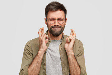 Wall Mural - Portrait of handsome unshaven guy presses lips, closes eyes, keeps fingers crossed, has strong believe in good luck before important trip, wills everything would be fine. God, help me please!