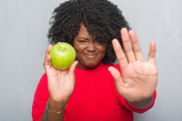 Poster - Young african american woman over grey grunge wall eating green apple with open hand doing stop sign with serious and confident expression, defense gesture