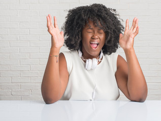 Poster - Young african american woman sitting on the table wearing headphones celebrating mad and crazy for success with arms raised and closed eyes screaming excited. Winner concept