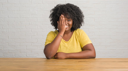 Canvas Print - Young african american woman sitting on the table at home covering one eye with hand with confident smile on face and surprise emotion.