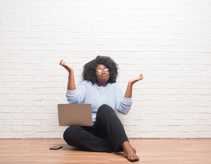 Poster - Young african american woman sitting on the floor using laptop at home clueless and confused expression with arms and hands raised. Doubt concept.