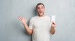 © Krakenimages.com - Young caucasian man over grey grunge wall showing notebook very happy and excited, winner expression celebrating victory screaming with big smile and raised hands