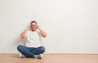© Krakenimages.com - Young caucasian man sitting on the floor over white brick wall covering ears with fingers with annoyed expression for the noise of loud music. Deaf concept.
