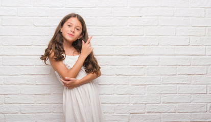 Wall Mural - Young hispanic kid over white brick wall with a big smile on face, pointing with hand and finger to the side looking at the camera.