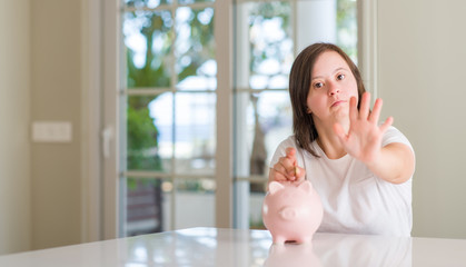 Sticker - Down syndrome woman at home holding piggy bank with open hand doing stop sign with serious and confident expression, defense gesture