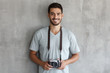 © Damir Khabirov - Smiling handsome male photographer in t shirt, holding retro film photo camera, standing against gray textured wall