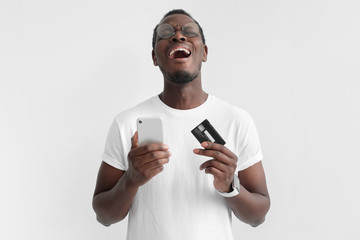 Transaction failed, declined. Young african american man holding credit card and smart phone in hand, crying, yelling because he has no cash, isolated on gray background