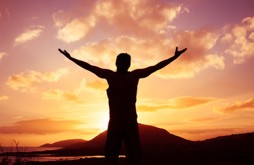 Feeling, happy and motivated. Young man with arms in the air on top of a mountain facing abeautiful sunset. 