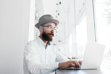 Bearded freelancer hipster wearing white shirt, stylish eyeglasses using laptop computer, typing on keyboard in modern office. Copywriter working a project, blog posts, email marketing at workplace.