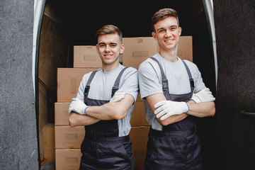 Wall Mural - Two young handsome smiling workers wearing uniforms are standing in front of the van full of boxes. House move, mover service.