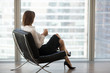 Successful businesswoman sitting in comfortable chair looking out of big window at the city, female boss or rich business lady relaxing enjoying view and morning coffee in modern office or hotel