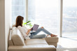 Relaxed woman using laptop in luxury home living room with big window, enjoying working, internet shopping, checking social network, reading news or communicating online with computer sitting on sofa
