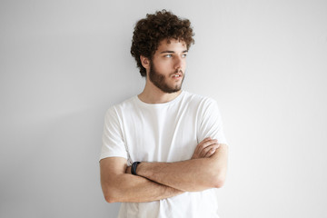 People and lifestyle concept. Portrait of fashionable young Caucasian bearded hipster wearing white t-shirt posing in studio with arms folded, looking away with serious thoughtful expression