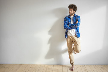 Isolated shot of attractive young Caucasian man with thick beard and bare feet posing indoors standing against white blank studio wall background with copy space for your promotional content