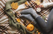 Women's Hands And Feet In Sweater And Woolen Cozy Gray Socks Holding Cup Of Hot Coffee With Marshmallow, Sitting On Plaid With Pumpkin, Knitted Scarf, Leaves. Concept Winter Comfort, Morning Drinking.