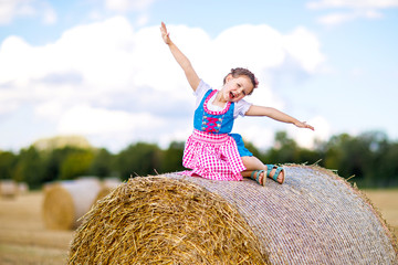 Wall Mural - Cute little kid girl in traditional Bavarian costume in wheat field. German child with hay bale during Oktoberfest in Munich. Preschool girl play at hay bales during summer harvest time in Germany.