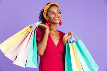 Poster - Image of happy african american woman 20s in hair band and earrings smiling and holding colorful paper packages, isolated over violet background