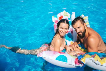 Wall Mural - Nice and young couple is leaning on air mattress and look on camera. They pose. Girl is smiling. Guy blink. They look happy.