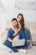 © LIGHTFIELD STUDIOS - smiling woman embracing boyfriend while he using laptop and sitting on floor at home