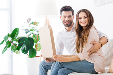 Poster - smiling couple with pizza in paper box sitting on sofa at home