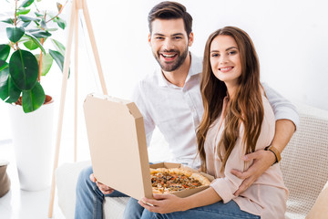 Poster - portrait of smiling couple with pizza in paper box sitting on sofa at home