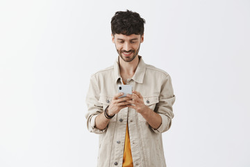Checking updates of favorite app in smartphone. Portrait of creative good-looking european man with beard and dark hair in trendy beige shirt over t-shirt typing message and smiling at device screen