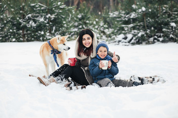  Little child and mom plays with funny Akita-inu dog in a winter park. Christmas happy family,mother and son walking with dog lying on snow in winter day. Drinking hot coffee or tea on snowy winter
