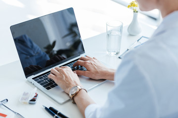 partial view of female doctor typing on laptop with blank screen at table in office