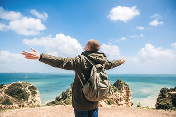 Wall Mural - View from the back. A young man tourist enjoys the beautiful views of the Atlantic Ocean and the landscape off the coast in Portugal and raises his hands upward showing how pleased he is.