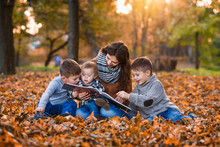 A Young Beautiful Mother Reads A Book With Fairy Tales For Her Three Sons In The Autumn Park. Mom With Children In The Autumn Background.