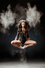 Wall Mural - beautiful ballerina in black bodysuit jumping on dark background with talc powder around
