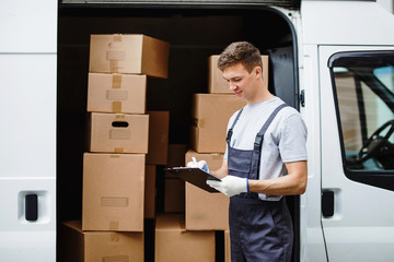 Wall Mural - A young handsome smiling worker wearing uniform is standing next to the van full of boxes holding a clipboard in his hands. House move, mover service.