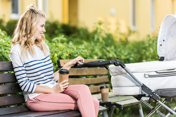 Wall Mural - side view of smiling mother sitting on bench with coffee to go and holding baby stroller in park