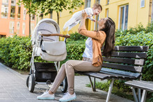 Side View Of Mother Holding Happy Baby On Bench Near Stroller
