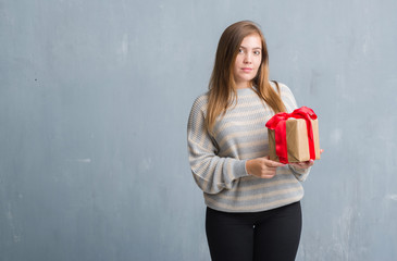 Wall Mural - Young adult woman over grey grunge wall holding a present with a confident expression on smart face thinking serious