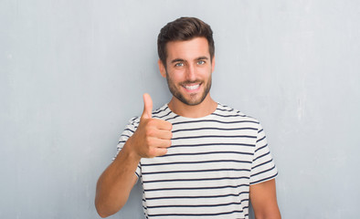 Handsome young man over grey grunge wall wearing navy t-shirt doing happy thumbs up gesture with hand. Approving expression looking at the camera with showing success.