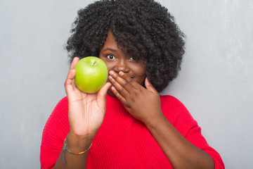 Canvas Print - Young african american woman over grey grunge wall eating green apple cover mouth with hand shocked with shame for mistake, expression of fear, scared in silence, secret concept