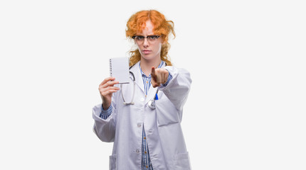 Sticker - Young redhead doctor woman holding blank notebook pointing with finger to the camera and to you, hand sign, positive and confident gesture from the front