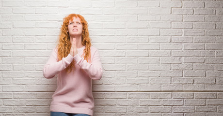 Sticker - Young redhead woman standing over brick wall begging and praying with hands together with hope expression on face very emotional and worried. Asking for forgiveness. Religion concept.